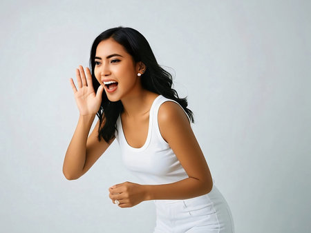 Cheerful young Asian woman shouting or announcing with hands near face, expressing excitement or surprise. Isolated on gray studio backgroundの素材