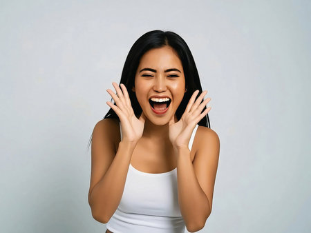 Cheerful young Asian woman shouting or announcing with hands near face, expressing excitement or surprise. Isolated on gray studio backgroundの素材
