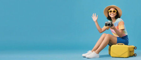 Happy young woman wearing sunglasses and hat, sitting on a suitcase with a camera, ready for vacation. Travel and holiday concept with cheerful tourist in bright colors on blue background.の素材