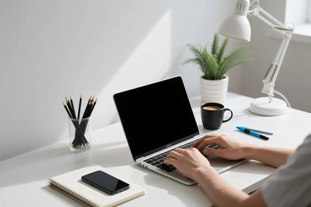 Modern minimalist workspace with laptop, notebook, smartphone, black coffee, lamp, pencils, and plant on white desk near window. Perfect for remote work, freelance, productivity, and creativity themes.の素材