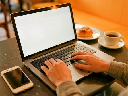 Young man working remotely from cafe, analyzing business growth chart on laptop while using smart phone. Cozy modern cafe setup with notebook and houseplant. Work from every where.の素材