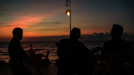 Silhouetted figures stand on a patio overlooking the ocean at sunset, creating a tranquil and...