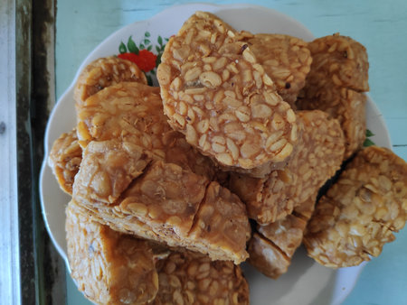 Fried tempeh sits piled high on a white plate, a delicious and savory snack ready to be enjoyed,...
