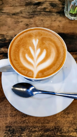 Cappuccino sits on white saucer with spoon on rustic wooden table in cozy cafe, creating a warm and inviting atmosphere.の写真素材