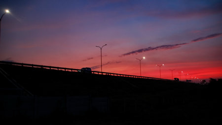 Crimson sky paints the horizon at dusk, casting a vibrant glow over the landscape with silhouette trees and streetlights, creating a dramatic and serene atmosphere.の写真素材