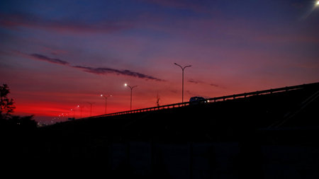 Crimson sky paints the horizon at dusk, casting a vibrant glow over the landscape with silhouette trees and streetlights, creating a dramatic and serene atmosphere.の写真素材
