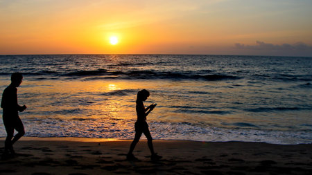 Couple walking along sandy beach at sunset, enjoying the peaceful atmosphere and the beautiful colors reflecting on the ocean, creating a romantic scene.の写真素材