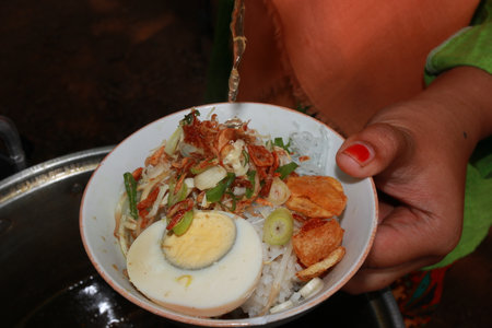 Hand pours broth into a bowl of soto ayam, a traditional Indonesian chicken soup with rice, egg, and vegetables, creating a warm and comforting meal.の写真素材