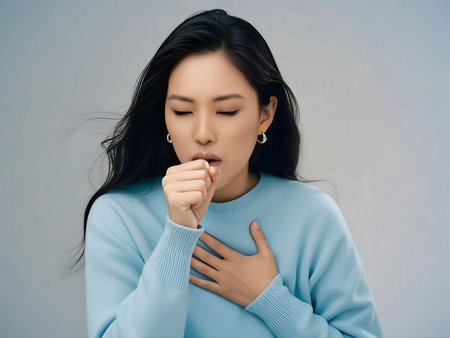 Young asian woman coughing while holding chest in indoor setting, feeling unwell and seeking relief from sickness.の素材