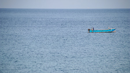 A blue sailing yacht travels across the horizon of the summer sea toward a sandy island beach under a clear ocean skyの写真素材