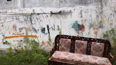 A discarded wooden sofa with red patterned upholstery lies on grassy ground against a peeling wall marked with graffiti, capturing a strong sense of abandonment and decay.の写真素材