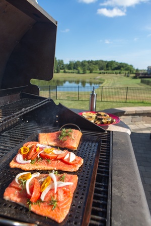 Fresh filleted salmon steaks on the barbecue topped with seasoning in the form of onion, tomato and herbs during preparation and grilling of a delicious seafood mealの写真素材