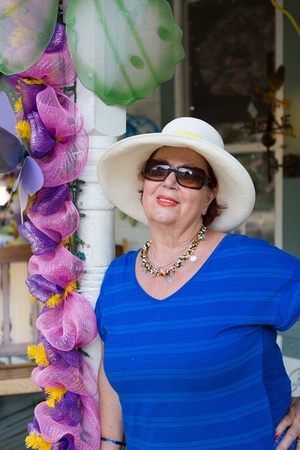 Stylish senior woman in sunglasses and a wide brimmed straw sunhat leaning against a pole alongside decorative pink flowersの写真素材