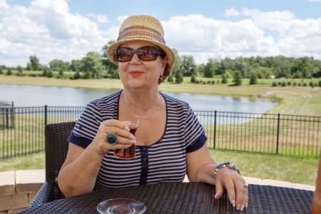 Stylish senior woman in a hat and sunglasses enjoying a drink outdoors sitting at a table overlooking a lakeの写真素材