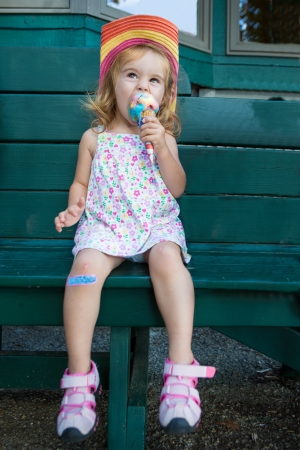 Cute little blond girl in a straw hat sitting on a wooden bench eating a colourful ice cream in the summer sunshineの写真素材