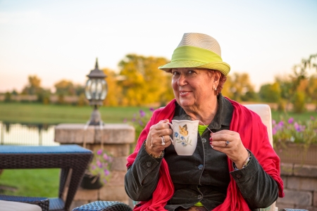 Senior woman with a straw green hat, sitting at the patio and showing her hot tea, perhaps she wants to stay warmの写真素材