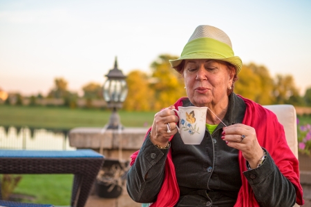 Senior woman with a straw green hat, sitting at the patio and blowing her hot tea, perhaps she wants to stay warmの写真素材