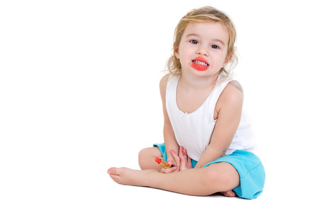 Cute very young girl practising to be a fashion model sitting on the floor grinning at the camera with her mouth smeared with bright red lipstick which she is holding in her hand, on whiteの写真素材
