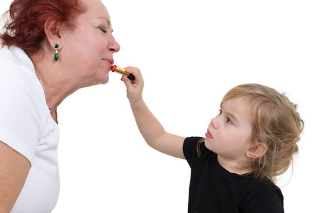 Young girl concentrates on applying lipstick to her grandmother's lips. Studio portrait over white.の写真素材