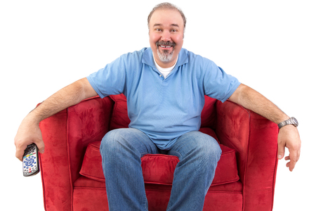 Funny cheerful middle-aged Caucasian man wearing casual clothes while sitting on a red comfortable armchair holding the remote control, on white backgroundの写真素材
