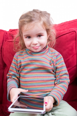 Beautiful toddler with tousled hair and an adorable smile sitting on a red sofa a tablet computer in her hands as she amuses herself at homeの写真素材