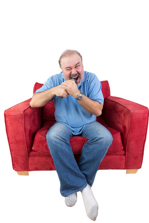 Frustrated middle-aged bearded man sitting in a red armchair biting a remote control from a television set screwing up his eyes in anger at a missed opportunity , isolated on whiteの写真素材