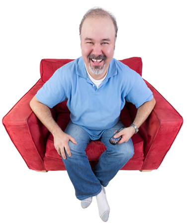 Middle-aged bearded man laughing as he watches television sitting in a comfortable armchair with the remote control in his handの写真素材