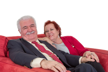 Smiling attractive fashionable senior couple relaxing hand in hand on a red sofa looking at the camera with happy smiles on a white backgroundの写真素材