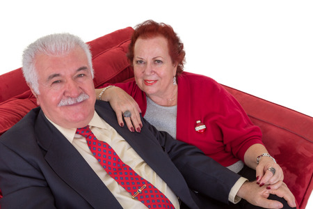 High angle view of an attractive stylish senior couple seated together on a red couch looking up at the camera with friendly smiles on a white backgroundの写真素材