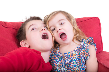 Two playful young children pulling faces as an attractive brother and sister sit together on a red couch playing copycat with their mouths wide openの写真素材