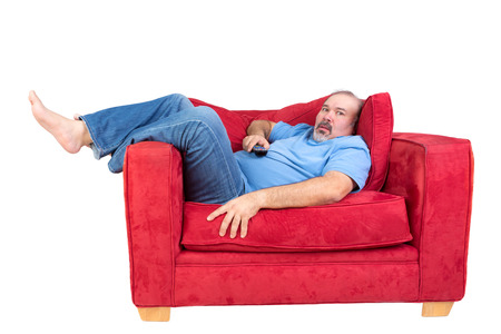 Man engrossed in watching television lying barefoot on a red couch with the remote control in his hand and a look of fascinated concentration, isolated on whiteの写真素材