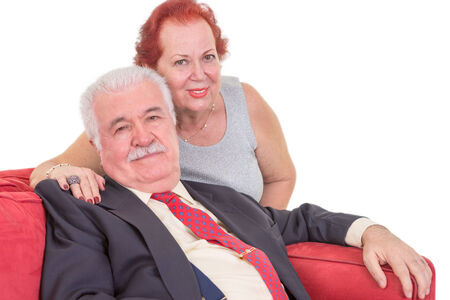 Loving elderly couple relaxing together on a red couch with the wife sitting behind her husband with her arm around his shoulders looking at the cameraの写真素材