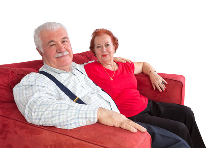 Attractive elderly couple smiling in satisfaction as they sit arm in arm on a comfortable red sofa over a white backgroundの写真素材