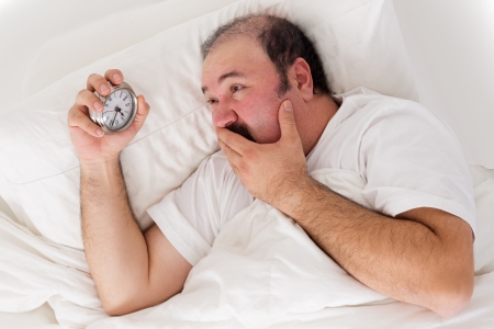 Man suffering from insomnia trying to sleep checking the time on his alarm clock in desperation as he realises he will not wake up in time for work if he sleeps nowの写真素材