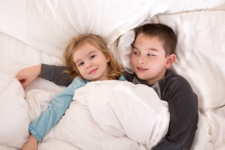Protective young boy lying in bed with his little sister watching over her as she lies smiling up at the camera as they prepare to go to sleepの写真素材
