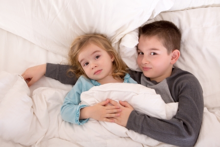 Affectionate little brother and sister lying in bed cuddling together under the duvet looking up sleepily at the cameraの写真素材