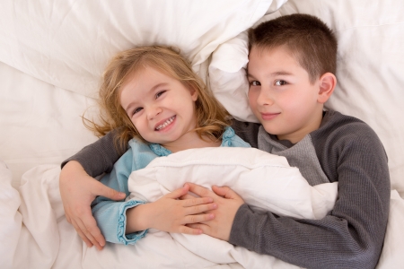 Mischievous young brother and sister in bed grinning up at the camera as they lie side by side under the duvet preparing to go to sleepの写真素材