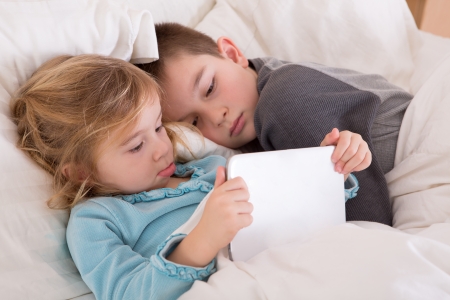 Cute little girl and boy reading a bedtime story as they lie together in bed looking at a tablet computer as they prepare to go to sleep with the little sister holding the notebookの写真素材