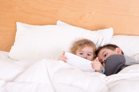 Little brother and sister snuggling up together at bedtime under a warm duvet as they read an e-book or surf the internet on a tablet-pcの写真素材
