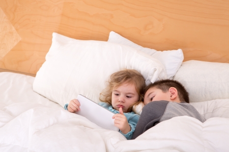 Tired little brother and sister snuggling up together in a warm comfortable bed as they read a bedtime story on a tablet computer before falling asleepの写真素材