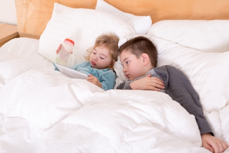 Happy little brother and sister lying in bed together as they cuddle under the duvet reading a tablet computer before going to sleepの写真素材
