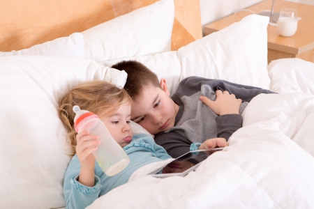 Tired little brother and sister going to sleep lying close together in a warm comfortable bed with a tablet computer on which they have just read a bedtime storyの写真素材