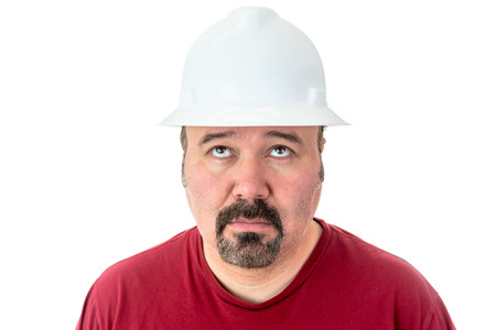 Glum looking workman wearing a hardhat looking for inspiration raising his eyes to the heavens in supplication, isolated on whiteの写真素材