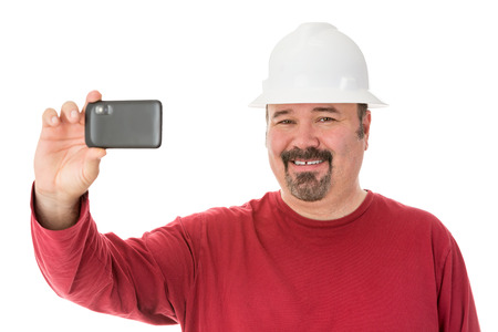 Smiling workman with a goatee beard wearing a hardhat taking a self-portrait using his smartphone,...