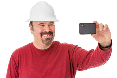 Workman with a neat goatee beard weaning a hardhat posing for a self-portrait giving a cheesy grin as he looks into the camera on his mobile phone, isolated on whiteの写真素材