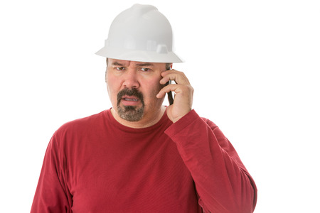Workman with a goatee beard wearing a hardhat listening to a call on his mobile phone with a look of shocked confusion isolated on whiteの写真素材