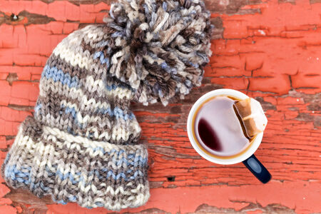 View from above of a cosy knitted woolly winter hat with a large pompom beside a cup of fresh hot tea on a grungy wooden surface with peeling red paintの写真素材