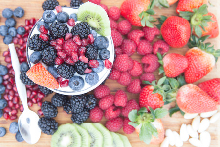 Healthy nutritious snack made of a bowl of raspberries, blackberries, blueberries, pomegranate, strawberries and kiwi, surrounded by fresh fruits on a wooden tableの写真素材