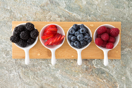 Assorted fresh berries in taster dishes viewed from above lined up on a wooden board on a stone kitchen counter with blackberries, sliced strawberry, blueberries and raspberriesの写真素材