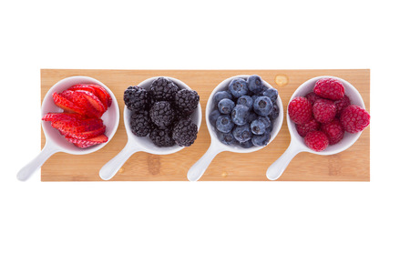 Variety of fresh ripe autumn berries served in small ceramic ramekins for a healthy refreshment of strawberries, raspberries, blueberries, and blackberries on a wooden board isolated on whiteの写真素材